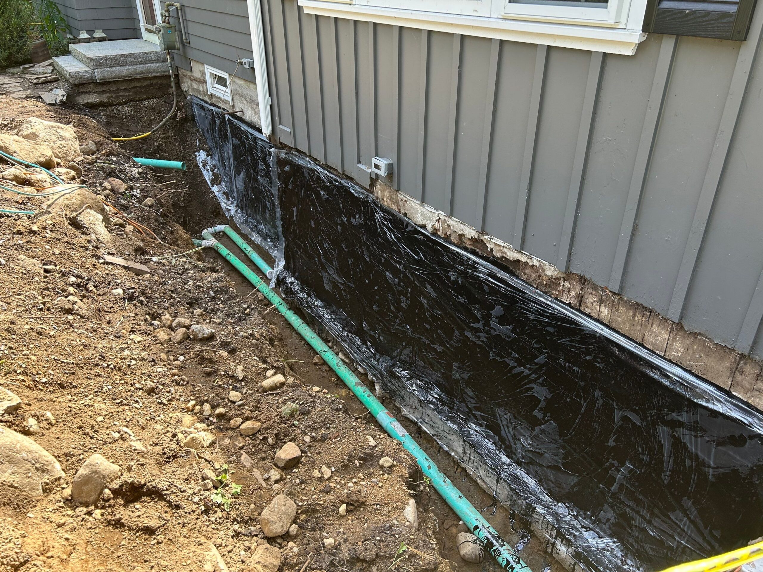 Water Runoff Solutions in DC, MD, VA - Our Services 1 Exterior wall foundation with plastic sheeting, drainage pipes, and exposed soil.