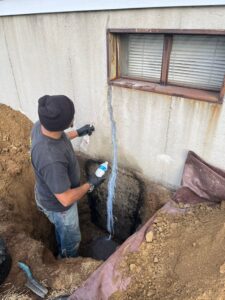 Worker repairing a vertical crack on an exterior foundation wall near a basement window.