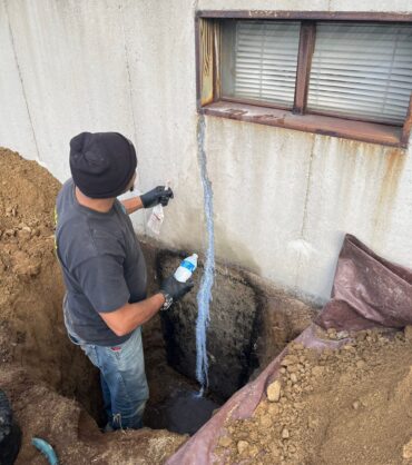 Worker repairing a vertical crack on an exterior foundation wall near a basement window.