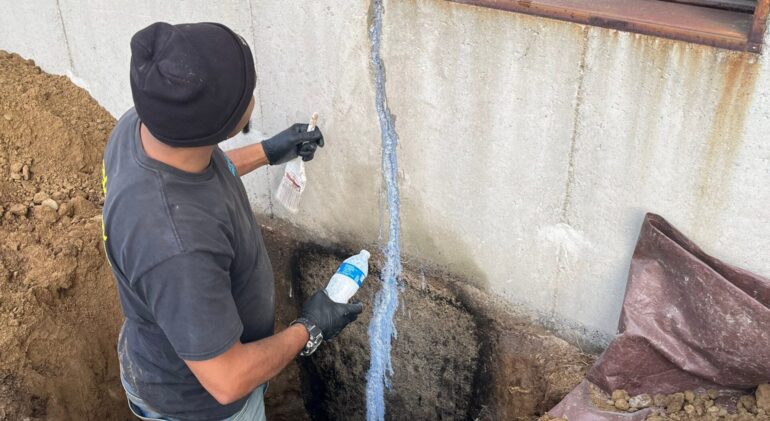 Worker repairing a vertical crack on an exterior foundation wall near a basement window.