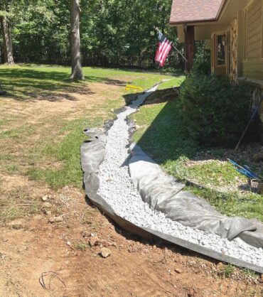 French drain installation with gravel-filled trench and drainage fabric along the side of a stone house.