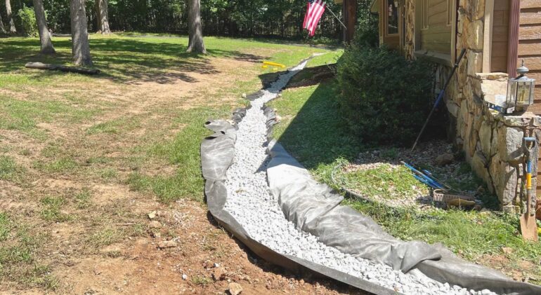 French drain installation with gravel-filled trench and drainage fabric along the side of a stone house.