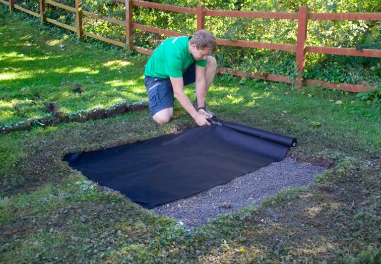 A person is laying down a black landscape fabric in a garden area as part of a drainage installation project.