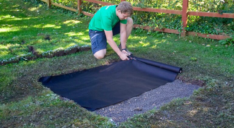 A person is laying down a black landscape fabric in a garden area as part of a drainage installation project.