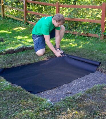 A person is laying down a black landscape fabric in a garden area as part of a drainage installation project.