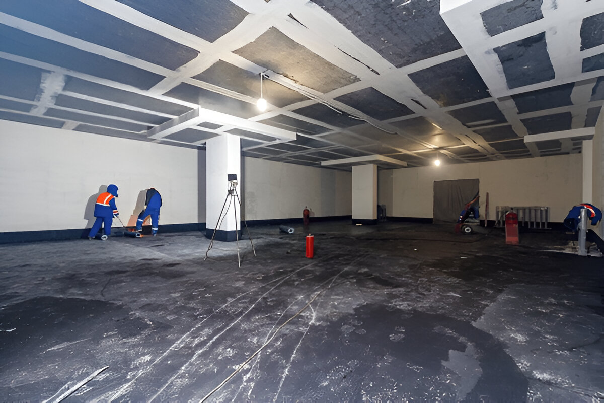 Workers applying waterproofing material on the floor of a large basement.