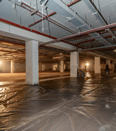 Construction workers installing plastic sheeting on the floor of a large underground basement.