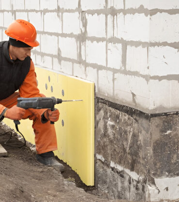 Worker drilling insulation boards into a building foundation wall.