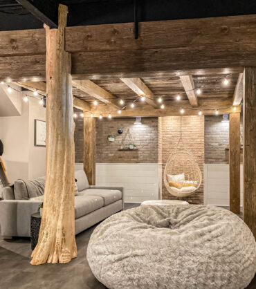 Rustic basement living room with exposed wooden beams, cozy seating, string lights, and a hanging chair for a warm, inviting atmosphere.