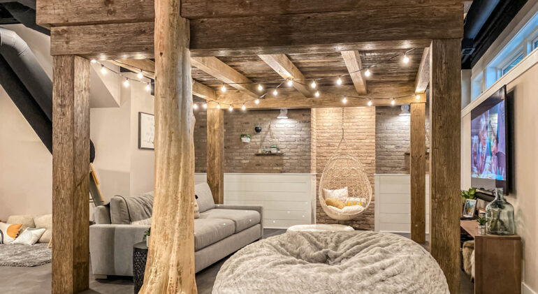 Rustic basement living room with exposed wooden beams, cozy seating, string lights, and a hanging chair for a warm, inviting atmosphere.