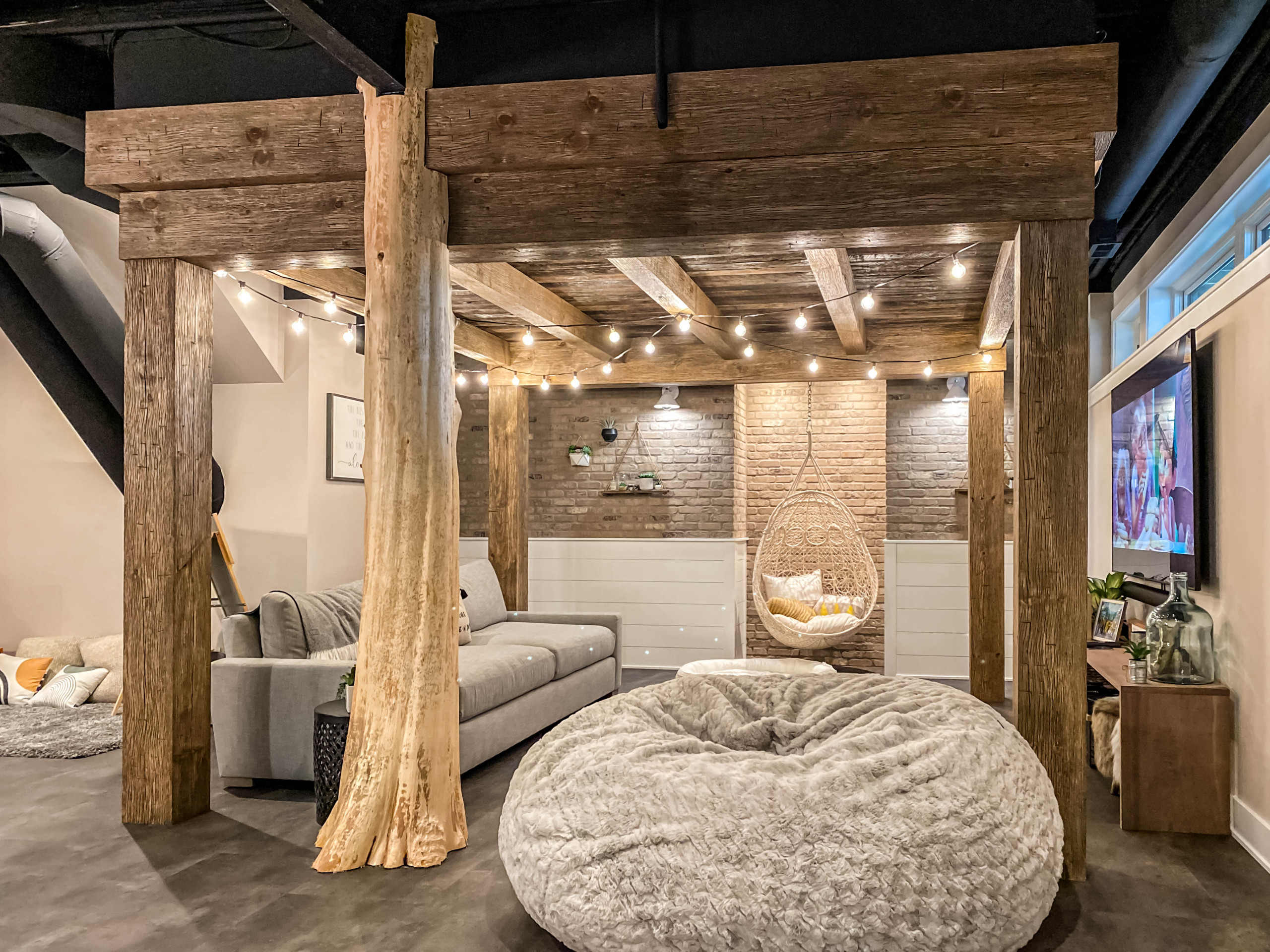 Rustic basement living room with exposed wooden beams, cozy seating, string lights, and a hanging chair for a warm, inviting atmosphere.