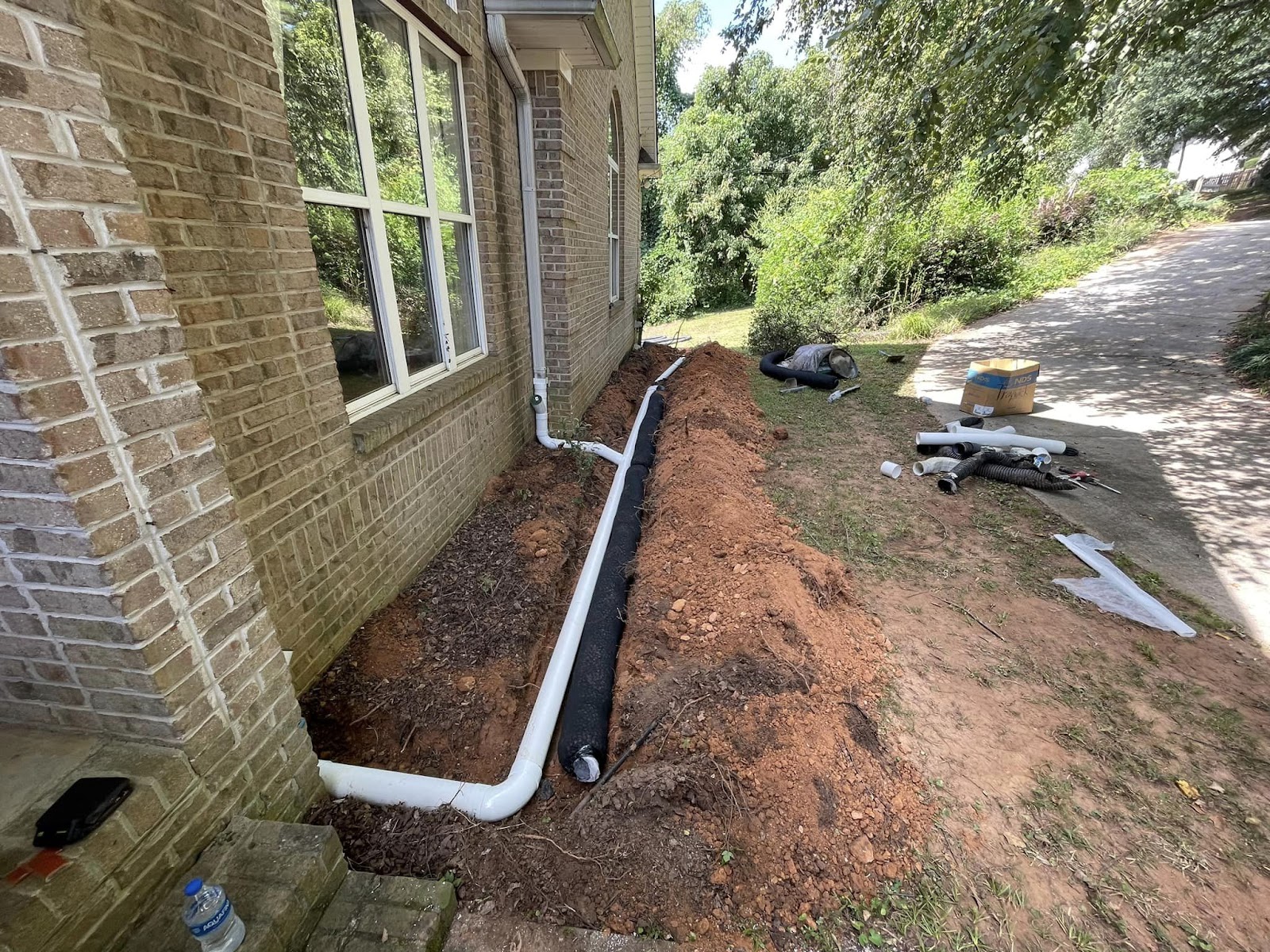 Exterior French drain system installation with white PVC pipes and drainage fabric along a brick house foundation.