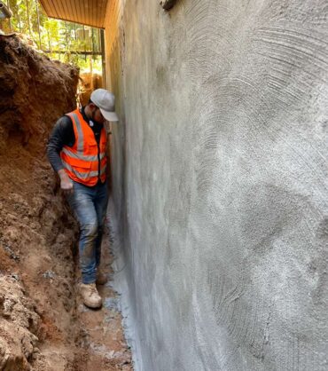 A DMV Waterproofing crew member in the excavated trench beside a foundation wall sealed with a cementitious waterproofing coat in Bethesda, MD