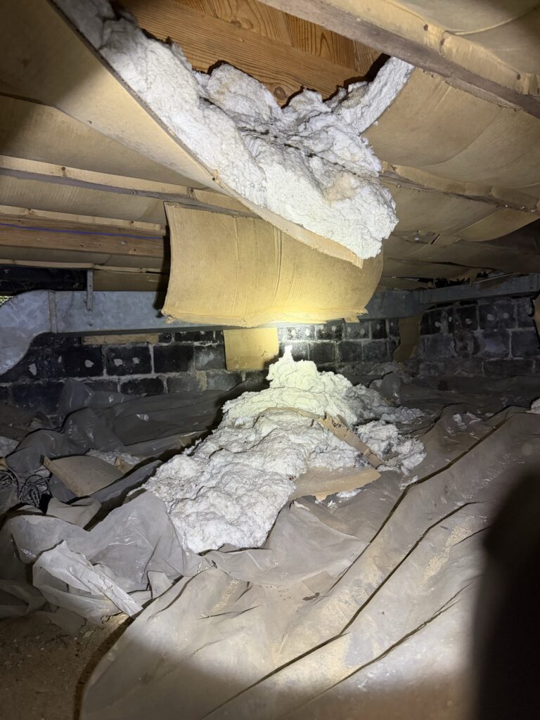 Fiberglass insulation batts hanging loose and fallen from crawl space floor joists onto an old plastic sheet below