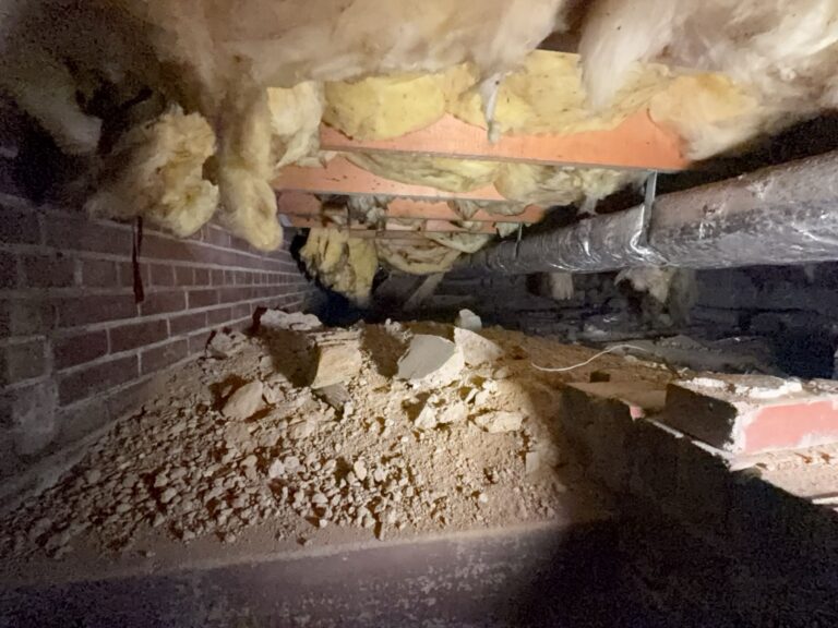 Fiberglass batts sagging out of the floor joists in a vented crawl space, over a bare dirt and rubble floor beside a brick foundation wall