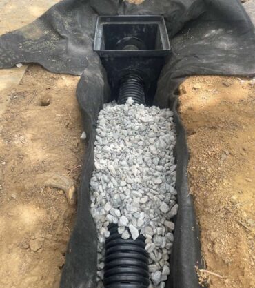 Catch basin box connected to corrugated drain pipe and surrounded by gravel inside a fabric-lined trench