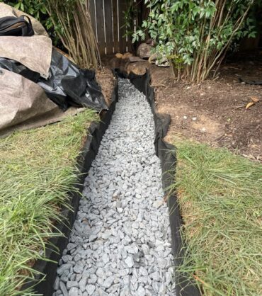 Completed drainage trench filled with washed gravel, geotextile fabric visible along both edges, running through a planted bed