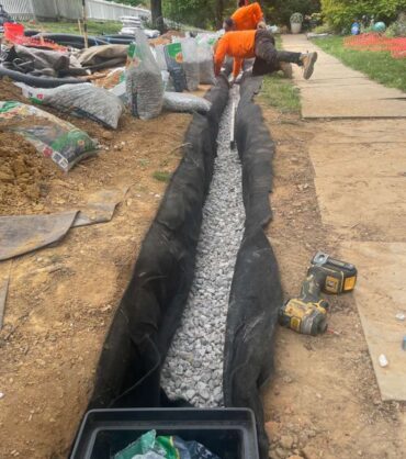 Straightedge laid along a gravel bed inside a fabric-lined drainage trench to verify consistent fall before the pipe goes in