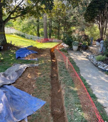 Long narrow drainage trench cut across a lawn beside a walkway, with excavated soil stacked on tarps alongside