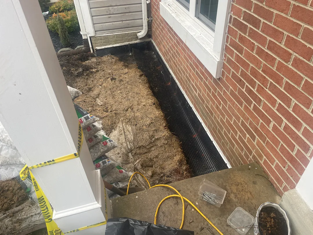 Exterior French Drain and Buried Downspout in Silver Spring, MD: A Real Front-Yard Project 1 Front planting bed opened for an exterior French drain in Silver Spring, with a dimpled drainage strip along the lower brick wall