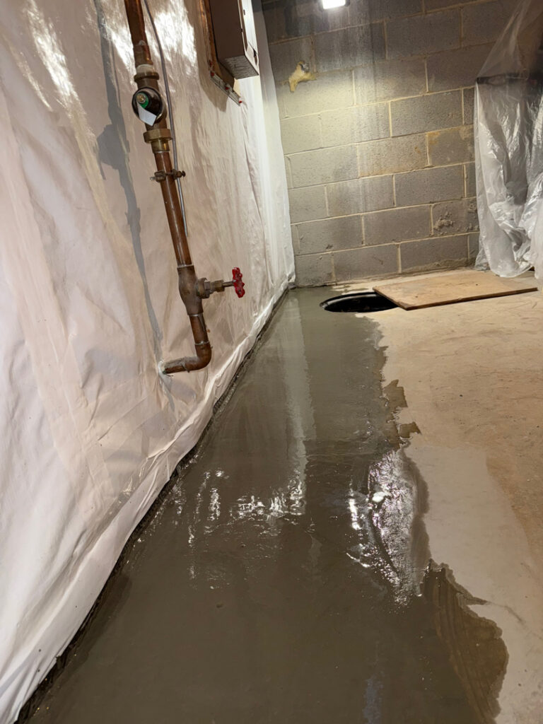 Fresh concrete beside a basement wall and open sump basin during interior drainage work
