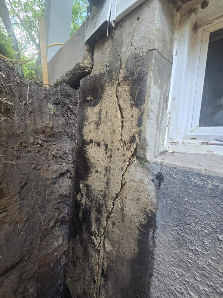 Exposed foundation wall with a visible vertical crack beside a basement window during exterior excavation