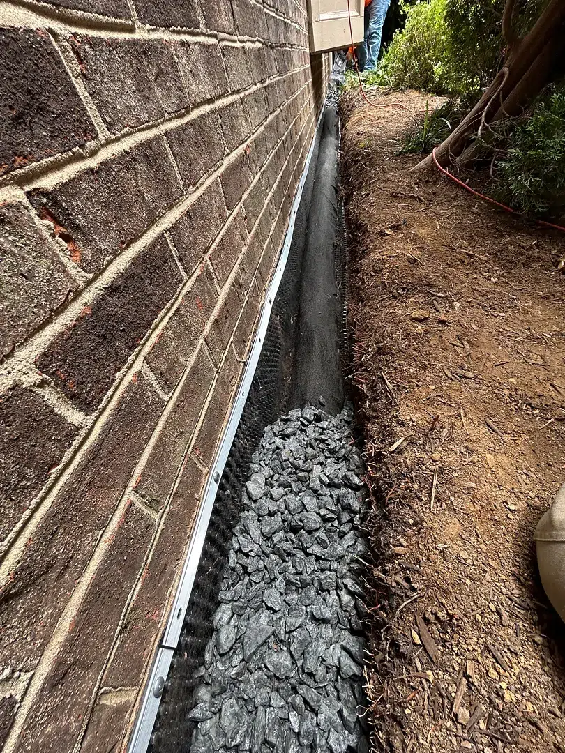 Gravel in a narrow exterior French drain trench beside a Rockville brick home